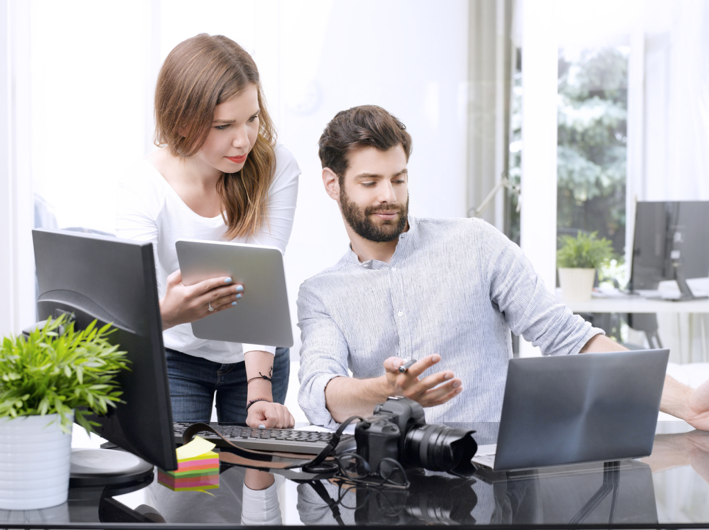 Portrait of young businessman and businesswoman working together online. Young professional sitting in front of laptop while his assistant standing next to him and holding hand digital tablet.